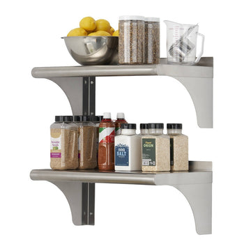 Two stainless steel shelves are mounted on a white wall, holding various kitchen items. The top shelf contains a metal bowl of lemons, two clear containers with dried goods, and a clear measuring cup with metal utensils, while the bottom shelf holds several spice jars and bottles.