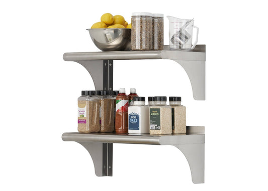 Two stainless steel shelves are mounted on a white wall, holding various kitchen items. The top shelf contains a metal bowl of lemons, two clear containers with dried goods, and a clear measuring cup with metal utensils, while the bottom shelf holds several spice jars and bottles.