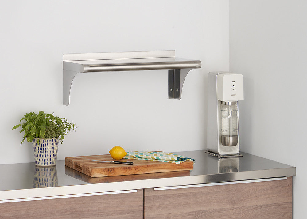 A stainless steel shelf is mounted on a white wall above a kitchen counter. The counter features a wooden cutting board with a yellow lemon and a knife, a potted green plant, and a white sparkling water maker.