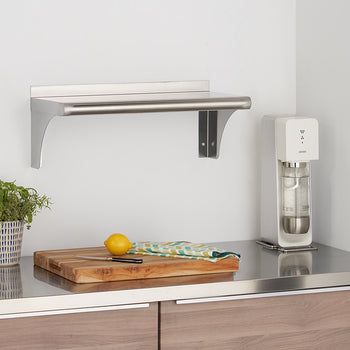 A stainless steel shelf is mounted on a white wall above a kitchen counter. The counter features a wooden cutting board with a yellow lemon and a knife, a potted green plant, and a white sparkling water maker.