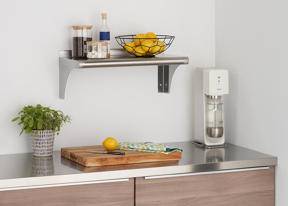 A stainless steel shelf is mounted on a white wall, holding two glass jars and a black wire basket filled with lemons. Below, a wooden cutting board rests on a stainless steel countertop next to a white sparkling water maker.