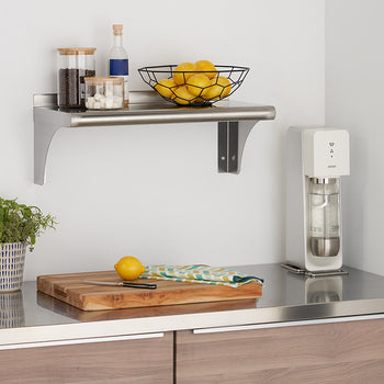 A stainless steel shelf is mounted on a white wall, holding two glass jars and a black wire basket filled with lemons. Below, a wooden cutting board rests on a stainless steel countertop next to a white sparkling water maker.