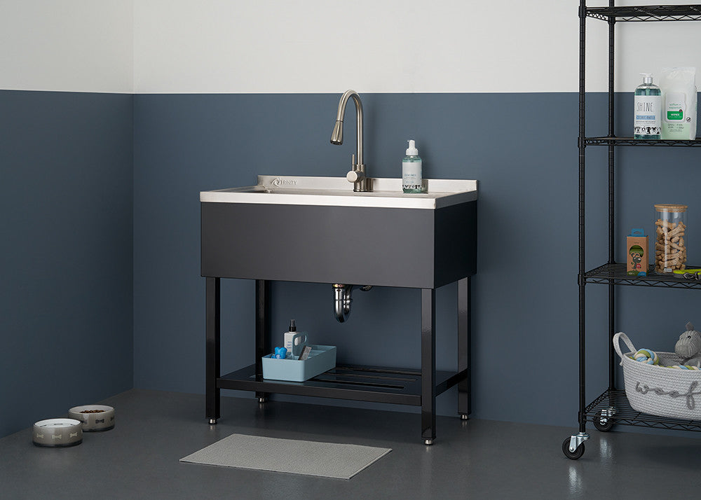 A stainless steel utility sink with a white basin and dark gray exterior is centered in the image, featuring a tall, curved faucet and a visible drain pipe. The sink is positioned on a black metal stand with a shelf, and is surrounded by a gray floor, two-tone gray walls, and a black wire shelving unit holding various cleaning supplies.
