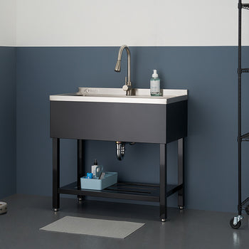 A stainless steel utility sink with a white basin and dark gray exterior is centered in the image, featuring a tall, curved faucet and a visible drain pipe. The sink is positioned on a black metal stand with a shelf, and is surrounded by a gray floor, two-tone gray walls, and a black wire shelving unit holding various cleaning supplies.