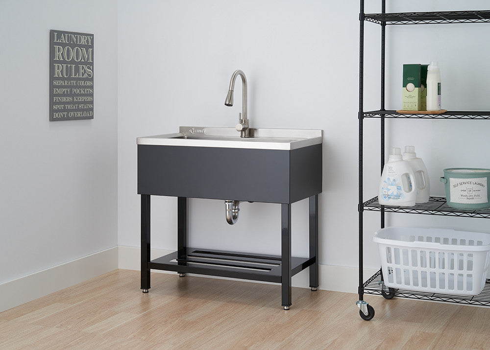 A dark gray utility sink with a stainless steel basin and a chrome faucet is centered in the frame, against a white wall with a framed sign. To the right, a black wire shelving unit on wheels holds various bottles and a white basket.
