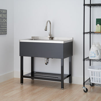 A dark gray utility sink with a stainless steel basin and a chrome faucet is centered in the frame, against a white wall with a framed sign. To the right, a black wire shelving unit on wheels holds various bottles and a white basket.