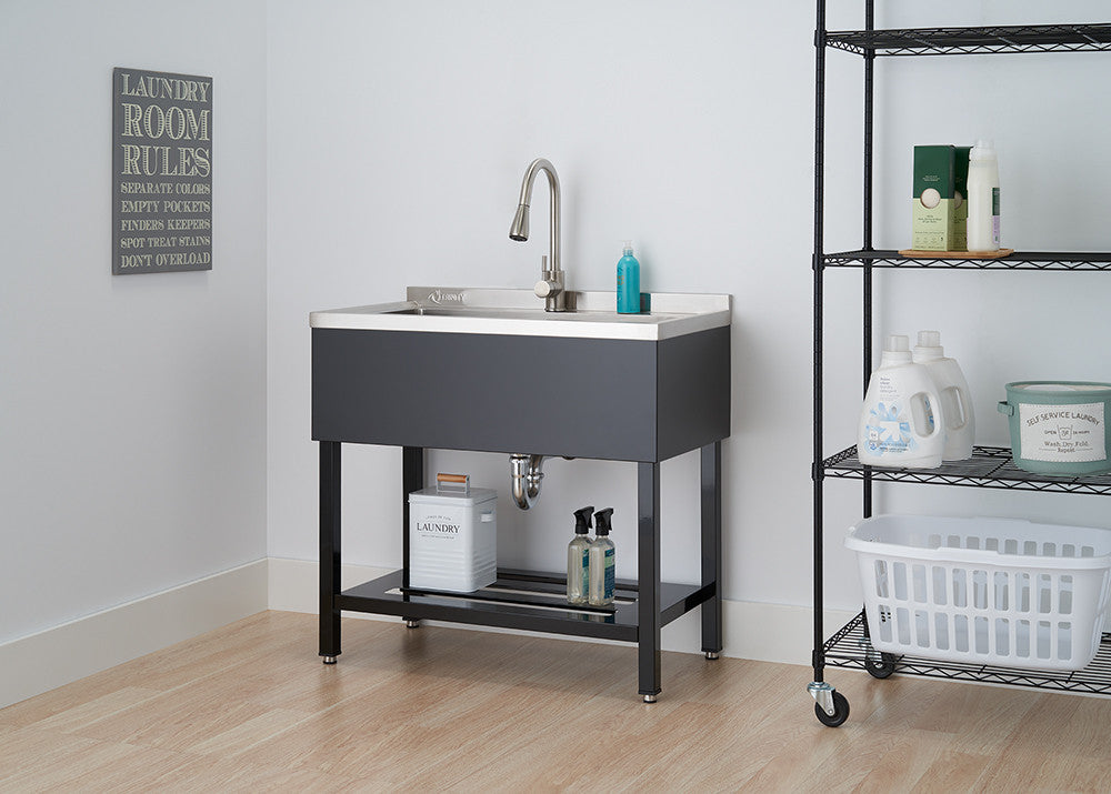 A freestanding utility sink with a stainless steel basin and dark gray cabinet is centered in the image. To the right, a black wire shelving unit holds cleaning supplies and a white laundry basket.
