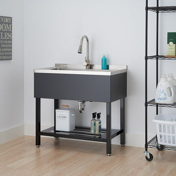 A freestanding utility sink with a stainless steel basin and dark gray cabinet is centered in the image. To the right, a black wire shelving unit holds cleaning supplies and a white laundry basket.