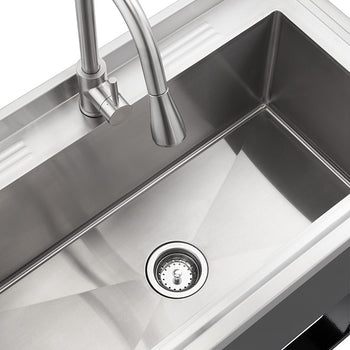 A close-up shot of a stainless steel utility sink with a curved faucet. The sink has a drain with a strainer, and the surrounding countertop is also stainless steel.