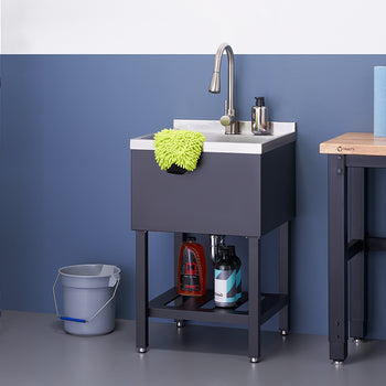 A black utility sink with a silver faucet stands in a garage or workshop setting. Below the sink, two bottles of cleaning solution sit on a shelf, and a grey bucket is placed next to the sink stand.