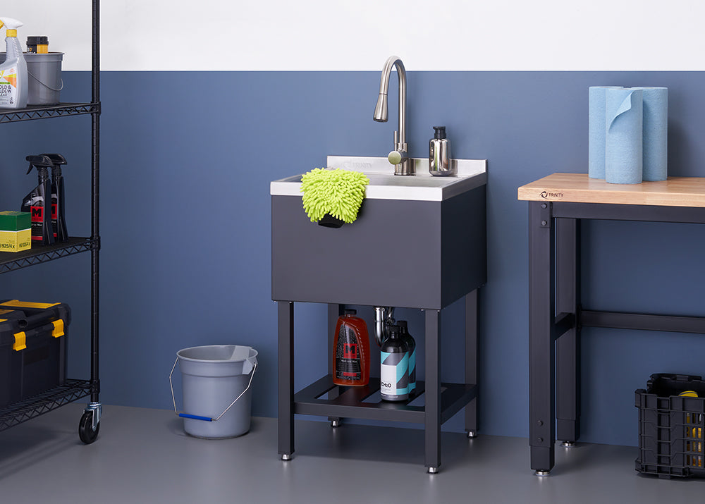 A black utility sink with a silver faucet stands in a garage or workshop setting. Below the sink, two bottles of cleaning solution sit on a shelf, and a grey bucket is placed next to the sink stand.