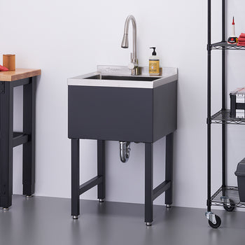 A modern utility sink with a stainless steel basin and dark gray cabinet stands on four legs against a white wall. A chrome faucet and soap dispenser are on the sink's edge, with a black wire shelving unit holding tools and supplies to its right.