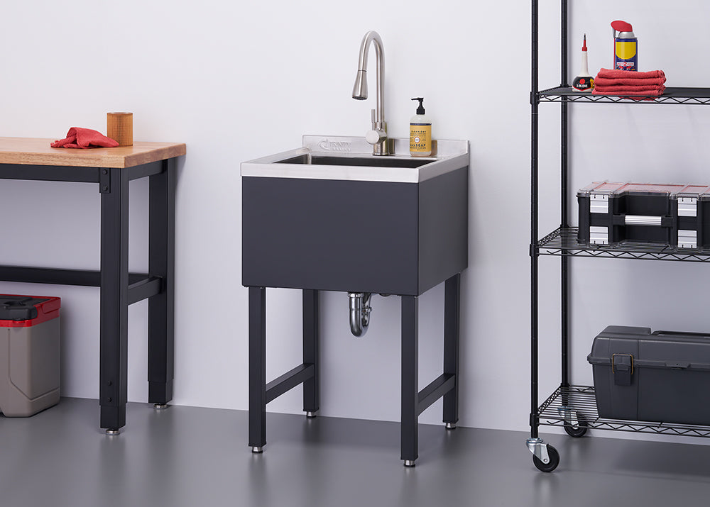 A modern utility sink with a stainless steel basin and dark gray cabinet stands on four legs against a white wall. A chrome faucet and soap dispenser are on the sink's edge, with a black wire shelving unit holding tools and supplies to its right.