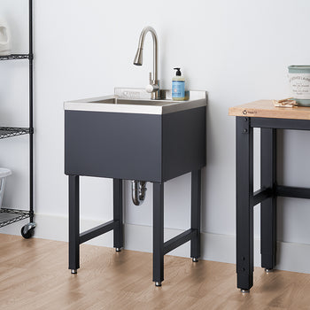 A stainless steel utility sink with a brushed nickel faucet stands on black metal legs against a white wall. To its left, a black wire shelving unit holds laundry supplies, and to its right, a wooden-topped workbench with black legs is partially visible.