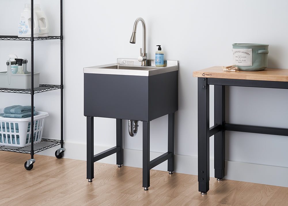 A stainless steel utility sink with a brushed nickel faucet stands on black metal legs against a white wall. To its left, a black wire shelving unit holds laundry supplies, and to its right, a wooden-topped workbench with black legs is partially visible.