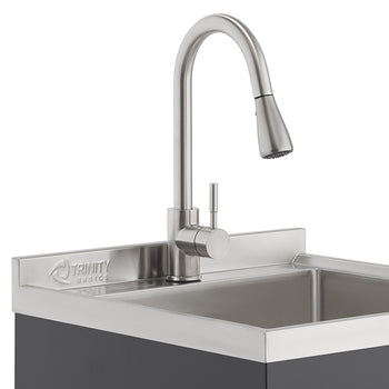 A close-up shot shows a stainless steel Trinity Basics sink with a modern, brushed nickel gooseneck faucet. The faucet features a pull-down sprayer head and a single handle control.