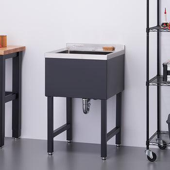 A modern utility sink with a black cabinet and stainless steel basin stands on four black legs. To its left is a wooden workbench, and to its right is a black wire shelving unit with various items.