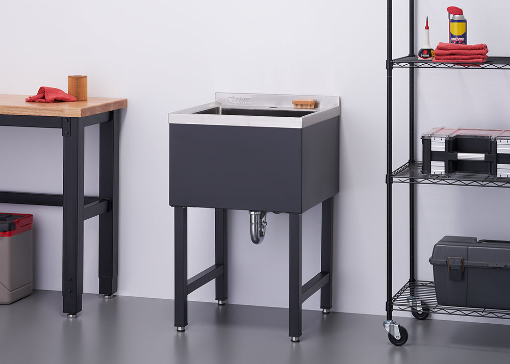 A modern utility sink with a black cabinet and stainless steel basin stands on four black legs. To its left is a wooden workbench, and to its right is a black wire shelving unit with various items.