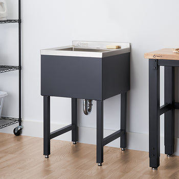 A laundry room features a black utility sink with a stainless steel top, positioned against a white wall. To the left is a black wire shelving unit on wheels with laundry supplies, and to the right, part of a wooden workbench with black legs is visible.