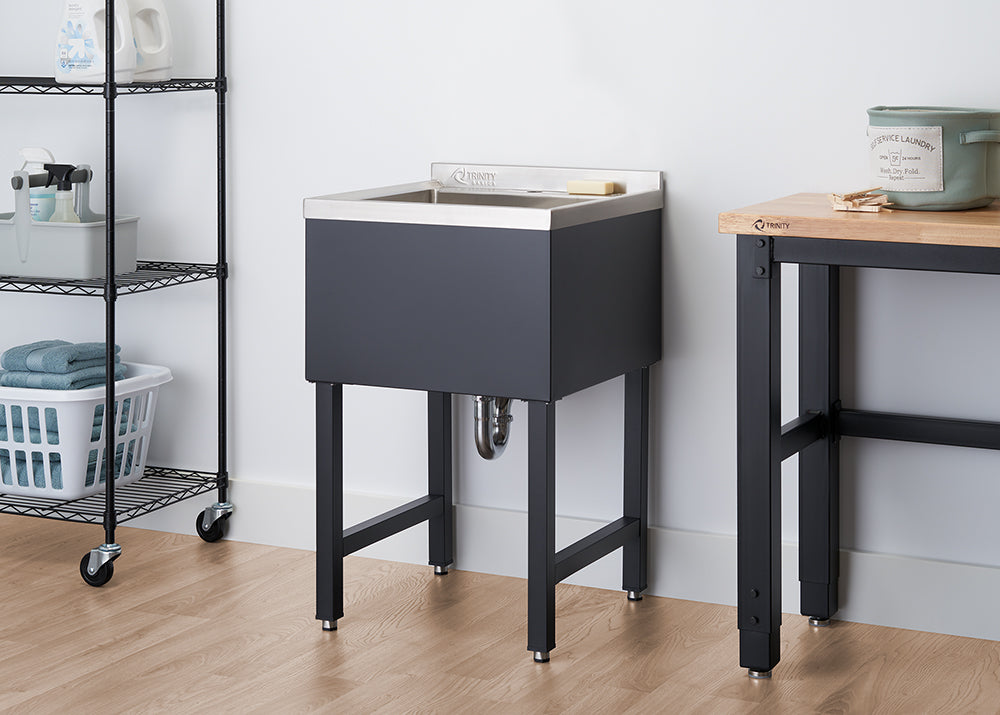 A laundry room features a black utility sink with a stainless steel top, positioned against a white wall. To the left is a black wire shelving unit on wheels with laundry supplies, and to the right, part of a wooden workbench with black legs is visible.