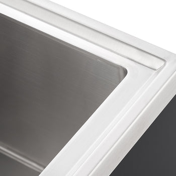 A close-up, high-angle shot shows the upper right corner of a modern, rectangular stainless steel sink. The brushed metal surface of the sink bowl and the flat edge around it are visible, with a dark gray counter or cabinet partially seen on the right.