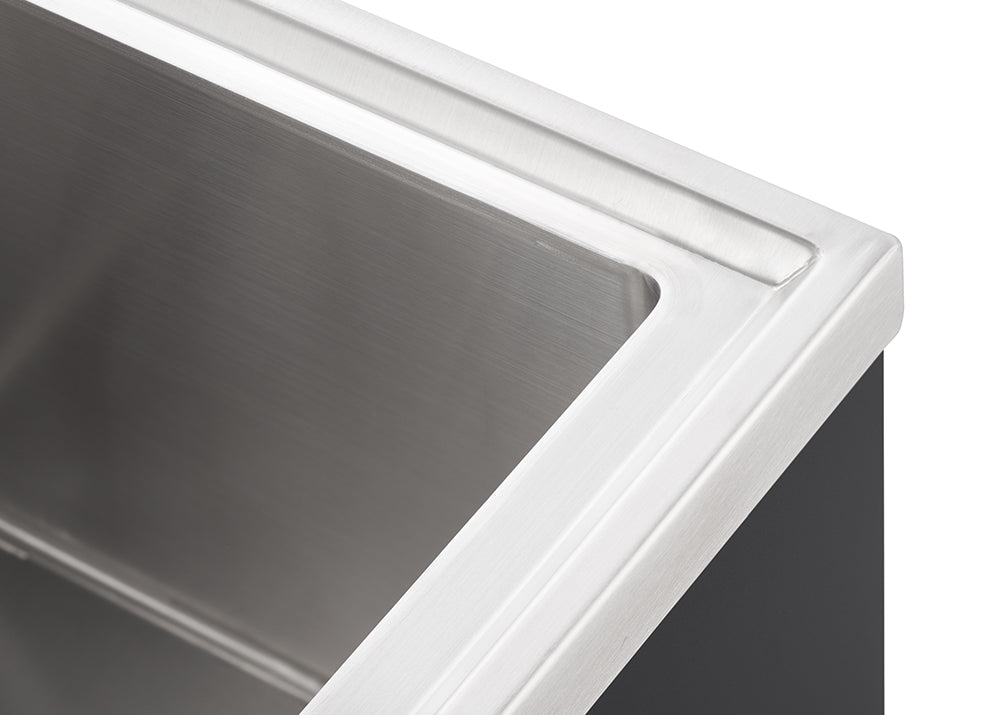 A close-up, high-angle shot shows the upper right corner of a modern, rectangular stainless steel sink. The brushed metal surface of the sink bowl and the flat edge around it are visible, with a dark gray counter or cabinet partially seen on the right.