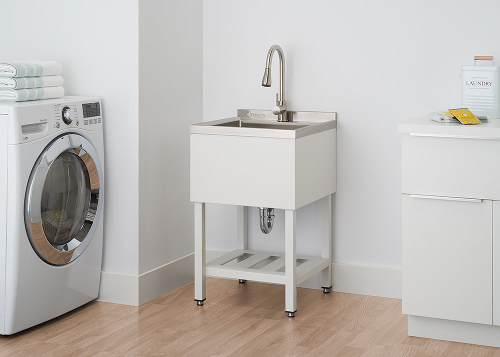 A modern white laundry room features a stainless steel utility sink with a white cabinet base and exposed silver legs. To the left, a large, silver front-loading washing machine sits next to a stack of folded towels.