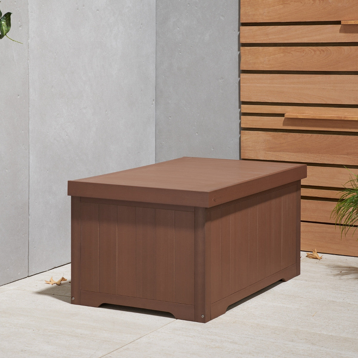 A brown, rectangular outdoor deck box with vertical paneling sits on a light-colored patio. The box has a flat, slightly overlapping lid and is positioned in a corner formed by a light gray wall and a horizontally paneled wooden wall.
