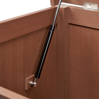 A close-up shows the interior of a brown outdoor deck box. A black gas strut is attached to the side wall with a metal bracket and extends upwards to support the open lid, which is partially visible at the top right.