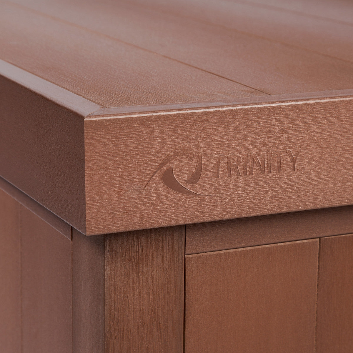 A close-up shot shows the corner and top of a brown outdoor deck box. The word "TRINITY" and a stylized wave design are embossed on the top panel.