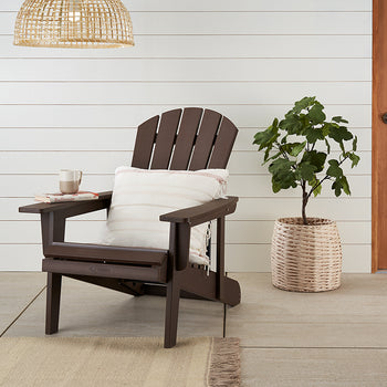 A dark brown Adirondack chair sits on a patio with a striped throw pillow and a mug on its armrest. Beside it, a potted plant in a woven basket and a light wooden door complete the outdoor scene.