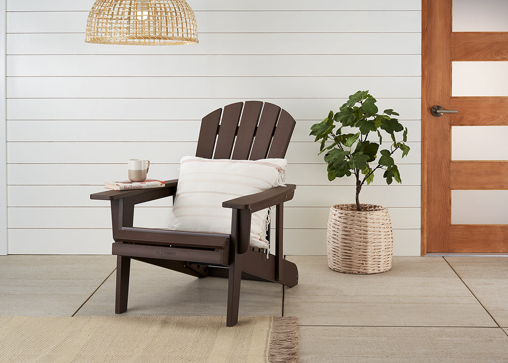 A dark brown Adirondack chair sits on a patio with a striped throw pillow and a mug on its armrest. Beside it, a potted plant in a woven basket and a light wooden door complete the outdoor scene.