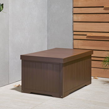 A rectangular, espresso brown outdoor storage box with a slightly lighter brown lid sits on a light-colored patio. To its left, green vines trail down a concrete wall, and to its right, a potted plant rests beside a wooden slatted wall.