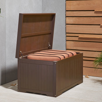 An espresso brown outdoor storage bench with its lid open, revealing a stack of orange cushions inside. The bench is situated on a light-colored patio next to a concrete wall with climbing green vines and a wooden slatted fence, with a potted plant on the right.