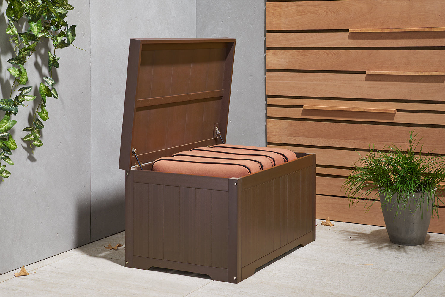 An espresso brown outdoor storage bench with its lid open, revealing a stack of orange cushions inside. The bench is situated on a light-colored patio next to a concrete wall with climbing green vines and a wooden slatted fence, with a potted plant on the right.