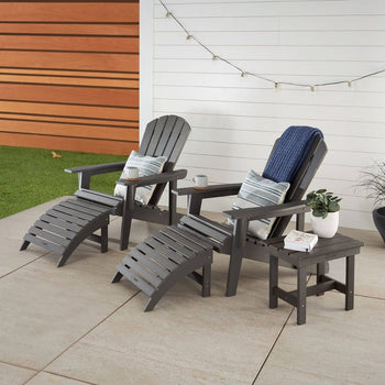 Two slate 5 piece gray Adirondack chairs with attached footrests sit on a patio, each adorned with a patterned pillow and a white mug on the armrest. A small matching side table holds a potted plant and a stack of white towels, while string lights hang overhead against a white plank wall.