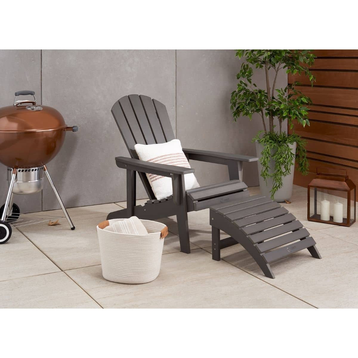 A slate gray Adirondack chair with an attached footrest and a striped white pillow sits on a patio. A copper-colored grill is visible to the left, and a white rope basket and a lantern with candles are on the tiled surface.