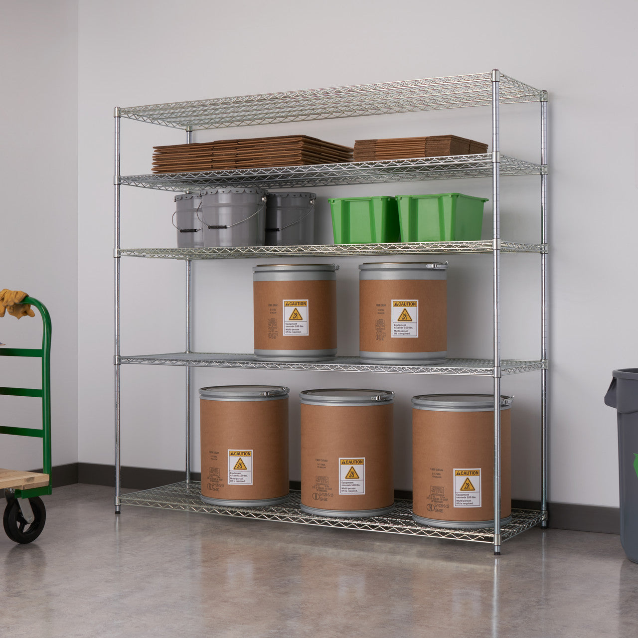 A silver, five-tier wire shelving unit stands against a light gray wall, holding various storage items. The shelves contain cardboard boxes, gray buckets, green bins, and large brown cylindrical containers with hazard labels.