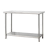 A stainless steel utility table with a rectangular top and four cylindrical legs is shown against a white background. The table also features a lower shelf for storage with trinity logo on the left side.