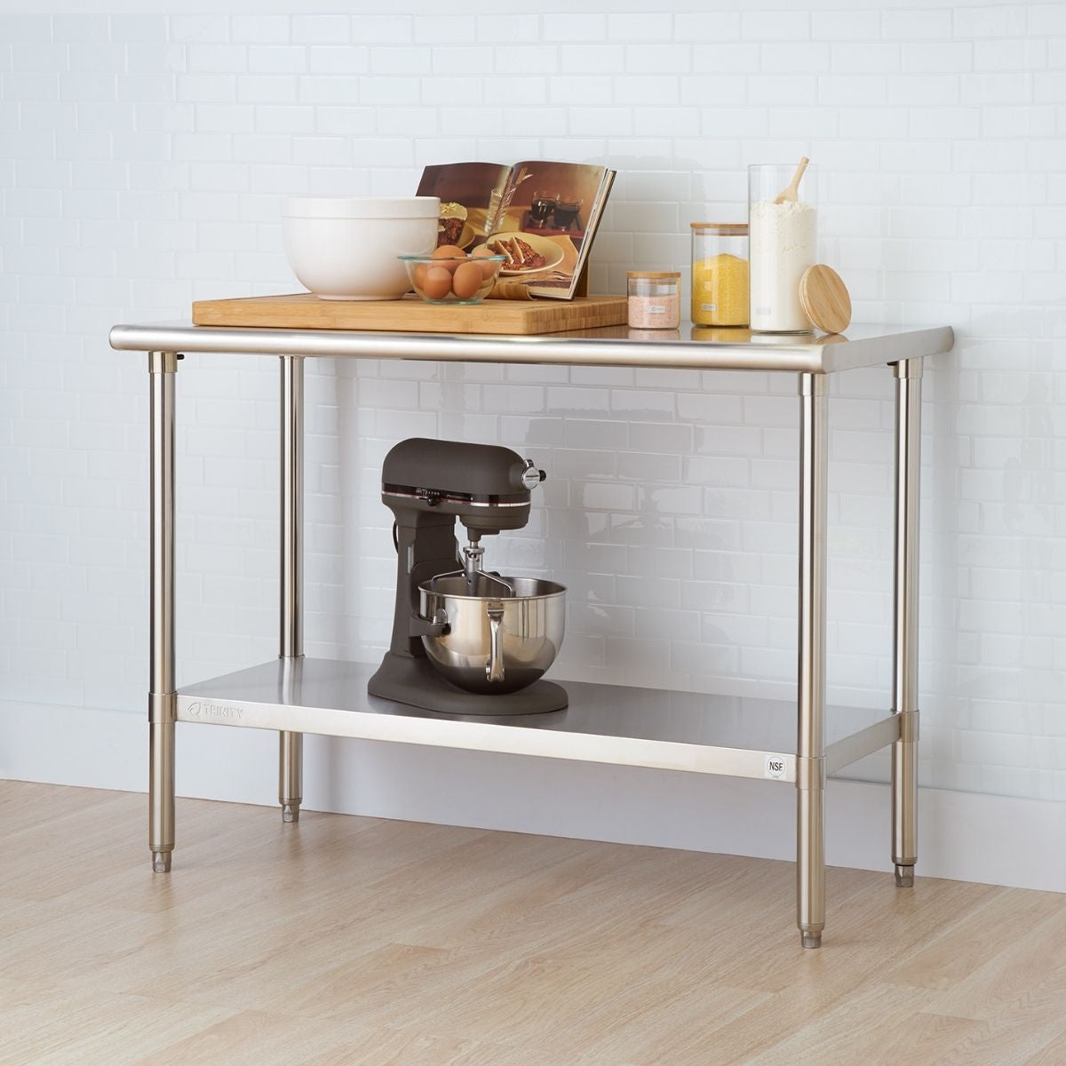 A stainless steel kitchen prep table stands against a white tiled wall. The table has two shelves and holds various kitchen items including bowls, eggs, a cutting board, flour, sugar, and a stand mixer.