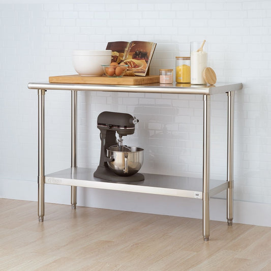 A stainless steel kitchen prep table stands against a white tiled wall. The table has two shelves and holds various kitchen items including bowls, eggs, a cutting board, flour, sugar, and a stand mixer.