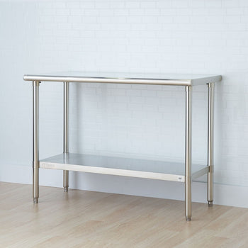A stainless steel utility table with a top surface, a lower shelf, and cylindrical legs stands against a white tiled wall and light wood-look flooring. The table appears to be a Trinity EcoStorage 48