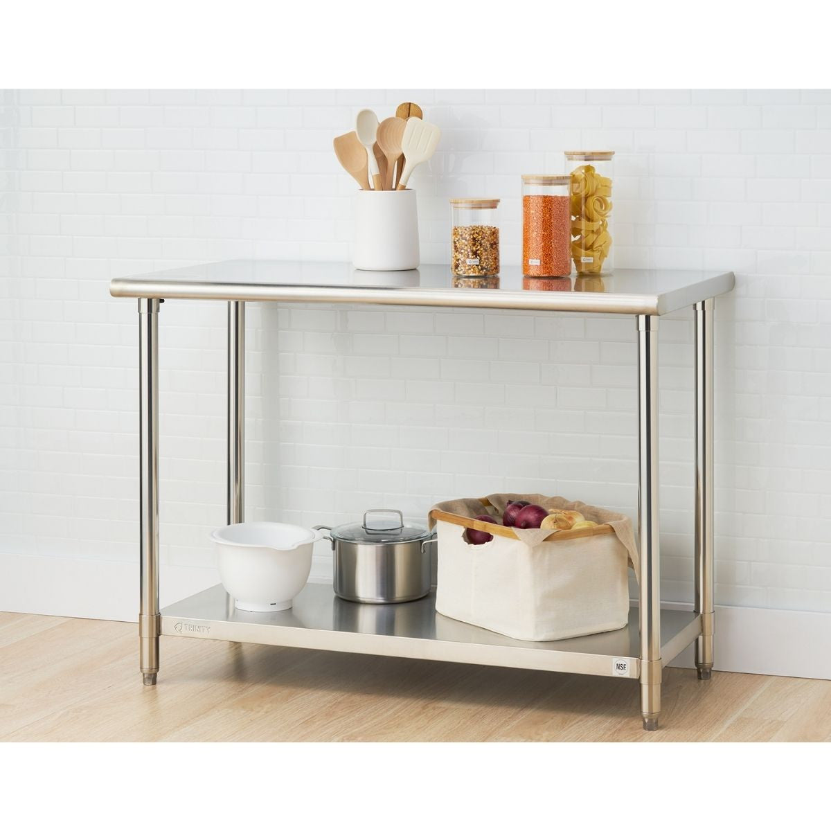 A stainless steel prep table with two shelves stands against a white tiled wall. The top shelf holds cooking utensils in a white holder and three glass jars filled with food, while the bottom shelf contains a white mixing bowl, a stainless steel pot with a lid, and a beige canvas bag filled with produce.