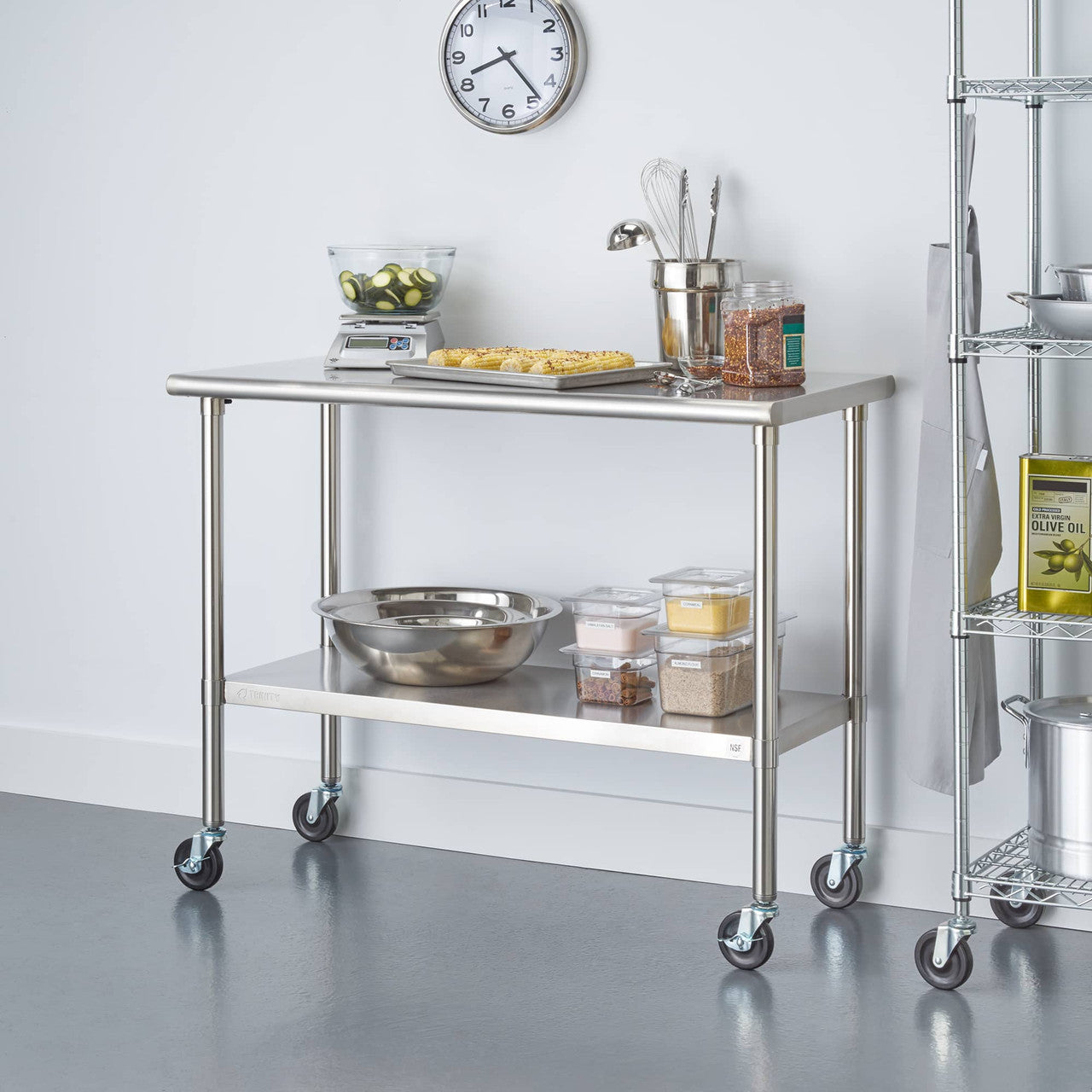 A stainless steel prep table with wheels holds kitchen items like a scale, a bowl of sliced cucumbers, a baking sheet with food, and various containers. The cart has two shelves and is positioned in a kitchen setting with a clock on the wall and part of a wire shelving unit visible on the right.