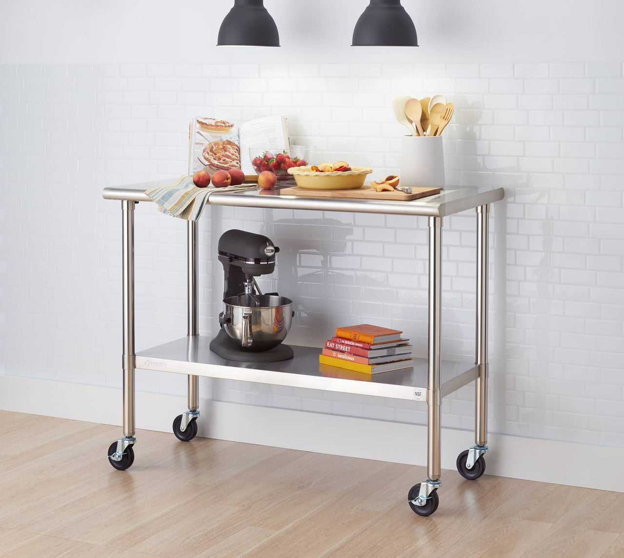 A stainless steel kitchen prep table with wheels sits on a light wood floor against a white brick wall. The table has two shelves and holds items such as a stand mixer, cookbooks, peaches, and a pie.