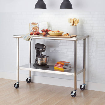 A stainless steel kitchen prep table with wheels sits on a light wood floor against a white brick wall. The table has two shelves and holds items such as a stand mixer, cookbooks, peaches, and a pie.