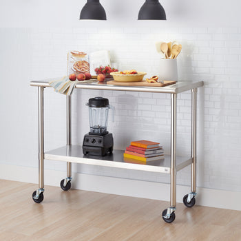 A stainless steel rolling kitchen prep table with wheels stands in front of a white subway tile wall. The tabletop is set with fresh peaches, strawberries, an open cookbook, a yellow pie dish, and wooden utensils, while a blender and a stack of colorful books sit on the bottom shelf.