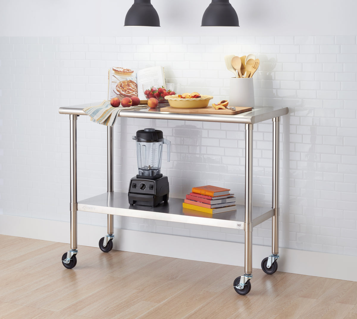 A stainless steel rolling kitchen prep table with wheels stands in front of a white subway tile wall. The tabletop is set with fresh peaches, strawberries, an open cookbook, a yellow pie dish, and wooden utensils, while a blender and a stack of colorful books sit on the bottom shelf.