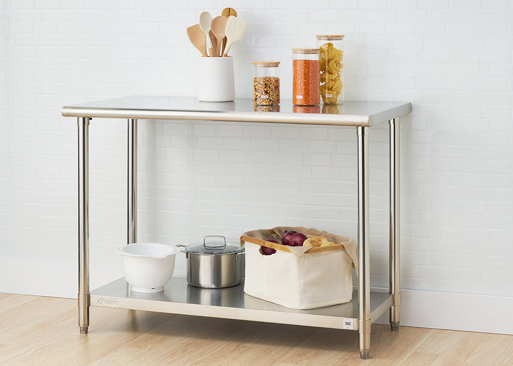 A stainless steel utility table with two shelves stands against a white tiled wall and a light wood floor. The top shelf holds wooden cooking utensils in a white container and three glass jars filled with dry goods, while the bottom shelf contains a white mixing bowl, a small stainless steel pot with a lid, and a canvas storage bin with red onions.