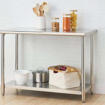 A stainless steel utility table with two shelves stands against a white tiled wall and a light wood floor. The top shelf holds wooden cooking utensils in a white container and three glass jars filled with dry goods, while the bottom shelf contains a white mixing bowl, a small stainless steel pot with a lid, and a canvas storage bin with red onions.
